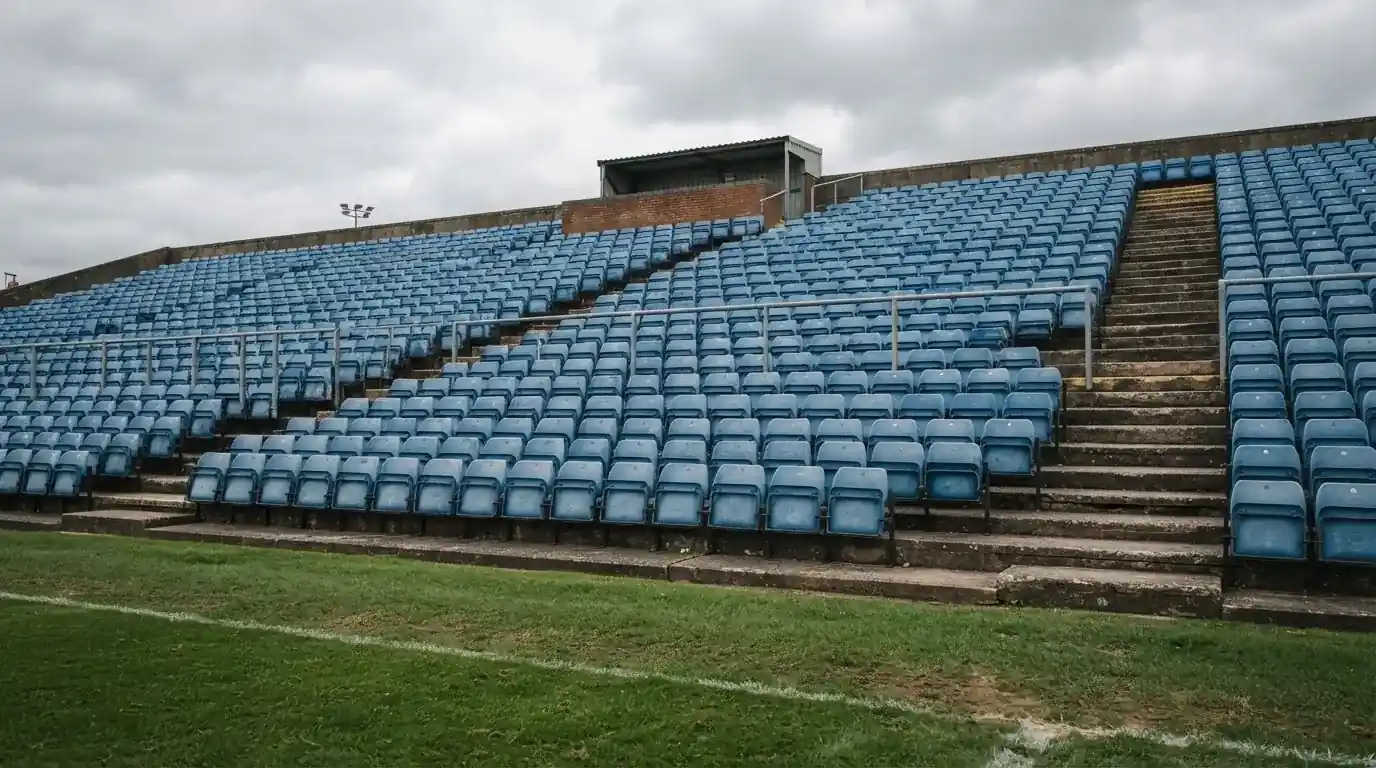 Rzędy pustych plastikowych krzesełek małego stadionu widziane z poziomu murawy pod pochmurnym niebem.