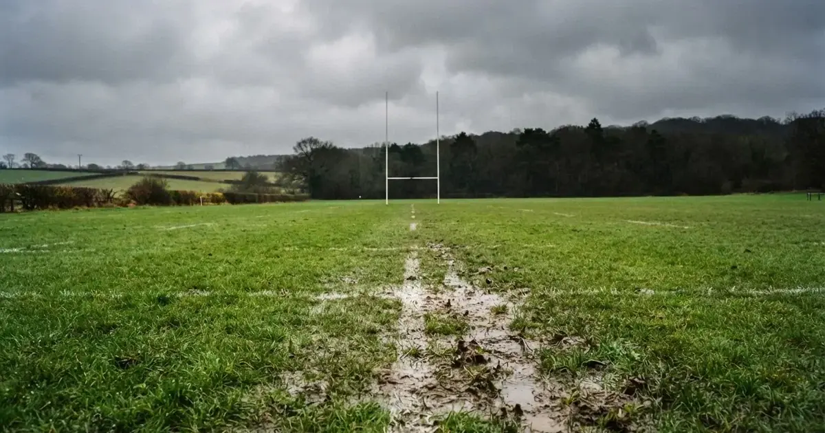 Boisko do rugby ze słupami bramki w oddali pod pochmurnym niebem, mokra trawa i ślady butów.